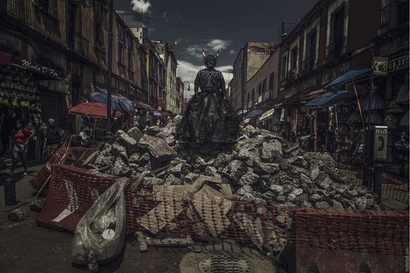 Cabaret Silencio standing atop a pile of rubble on a street in the Historic Center of Mexico City, wearing horns, a chain necklace, and a black skirt.